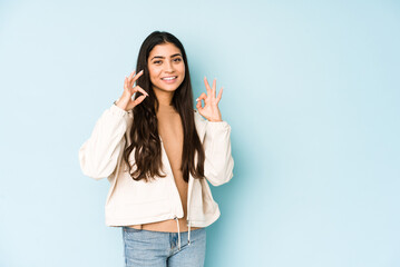 Young indian woman on blue background cheerful and confident showing ok gesture.