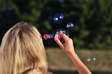 Blonde girl blowing soap bubbles outdoor in park on summer sunset. Happiness in nature concept....
