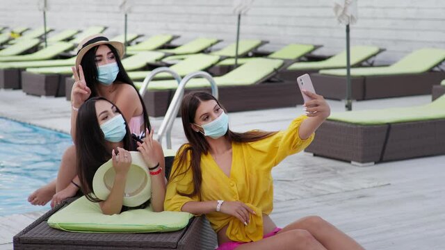 coronavirus vacation, adorable young women wear protective medical masks during social distance taking selfie by pool during summer holiday at resort with sun loungers in background