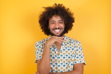 Young man with afro hair over wearing hawaiian shirt standing over yellow background Optimistic keeps hands partly crossed and hand under chin, looks at camera with pleasure. Happy emotions concept.