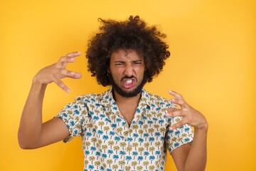 Young man with afro hair over wearing hawaiian shirt standing over yellow background Shouting frustrated with rage, hands trying to strangle, yelling mad.