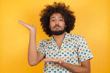 Young man with afro hair over wearing hawaiian shirt standing over yellow backgro pointing aside with both hands showing something strange and saying: I don't know what is this. Advertisement concept.