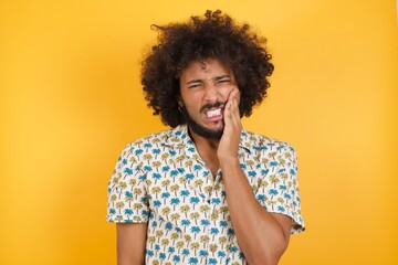 Young man with afro hair over wearing hawaiian shirt standing over yellow background touching mouth with hand with painful expression because of toothache or dental illness on teeth.