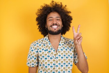 Young man with afro hair over wearing hawaiian shirt standing over yellow background showing and pointing up with fingers number two while smiling confident and happy.