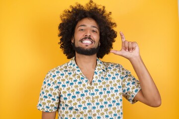 Young man with afro hair over wearing hawaiian shirt standing over yellow background smiling and confident gesturing with hand doing small size sign with fingers looking and the camera. Measure 