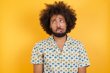 Fototapeta na wymiar Portrait of Young man with afro hair wearing hawaiian shirt standing over yellow wall making grimace and crazy face, screaming out of control, funny lunatic expressing freedom and wild.
