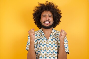 Young man with afro hair wearing hawaiian shirt standing over yellow wall being excited for success...