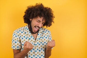 Young man with afro hair wearing hawaiian shirt standing over yellow wall very happy and excited...