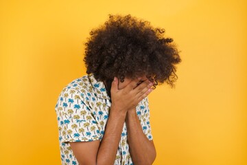 Sad Young man with afro hair wearing hawaiian shirt standing over yellow wall crying covering her face with her hands.