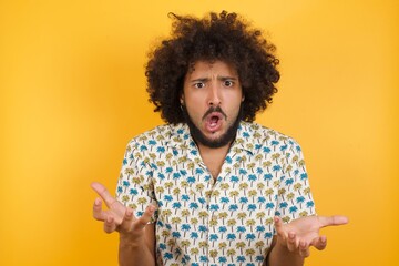 Frustrated Young man with afro hair wearing hawaiian shirt standing over yellow wall. feels puzzled and hesitant, shrugs shoulders in bewilderment, keeps mouth widely opened, doesn't know what to do.