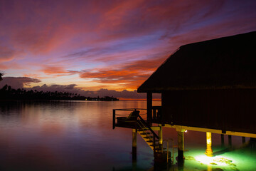 Overwater bungalow on a tropical island with a vibrant red sunrise in the sky over the ocean