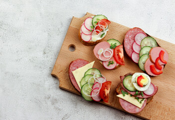 Assorted sandwiches with vegetables and sausages on the wooden cutting board on a light gray background. Top view, flat lay