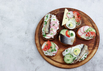 Assorted sandwiches with vegetables and fish on the round wooden cutting board on a light gray background. Top view, flat lay