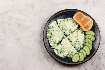 Scrambled eggs with green onions and cheese with toast and sliced cucumbers on a round plate on a light gray background. Top view, flat lay