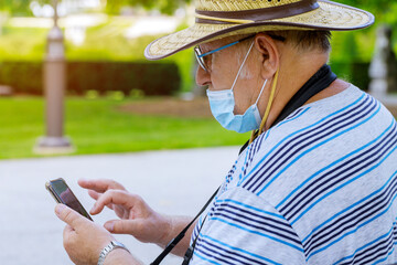 Portrait of an old man a wearing medical mask with using smartphone while listening protect during coronavirus in a park