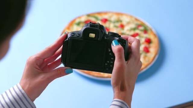  Top view - Female hands use dslr camera photographing appetizing fresh cooked italian pizza with tomatoes, cheese mozzarella, and sauce pesto on blue background. Food blogger of blogging concept. 