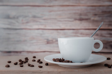 A white hot coffee cup and coffee beans with a spoon in the cup and a saucer is placed on a wooden plate and on a wooden background.