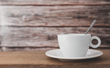 A white hot coffee cup with a spoon in the cup and a saucer is placed on a wooden plate and on a wooden background.