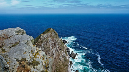 A narrow promontory juts out into the blue ocean. Waves crash against stones and foaming. There is a tiny house at the top of the cliff. Turquoise sky with clouds. Cape Point. South Africa.
