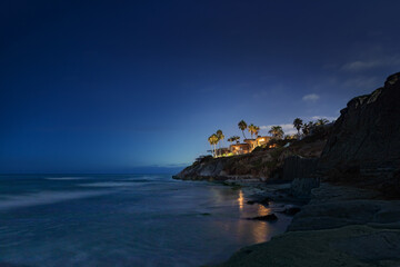 Clifftop homes overlooking the ocean at twilight with palm trees, lights reflecting in the water...
