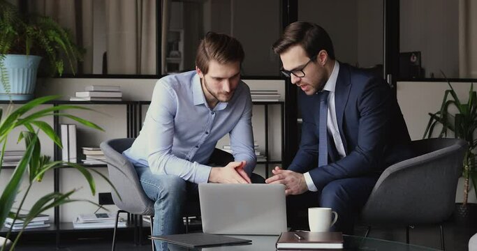 Two focused business partners looking at laptop screen, discussing corporate software application. Young male intern employee consulting about project with skilled boss leader at modern office.