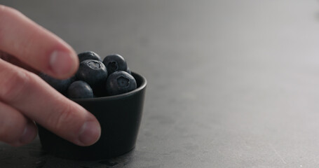 man hand take small black bowl with fresh blueberries on concrete surface with copy space