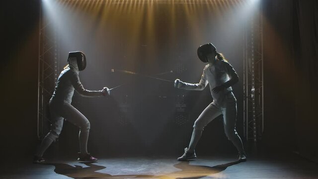 Side view duel of two women fencers. Sportswomen practice strikes, attack and defend. Filmed indoors with studio light. Slow motion.