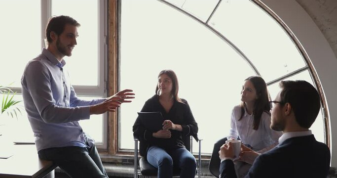 Young skilled business trainer leaning on table, sharing knowledge to attentive multiracial colleagues seated on chairs in office. Smart male financial expert educating interested coworkers indoors.