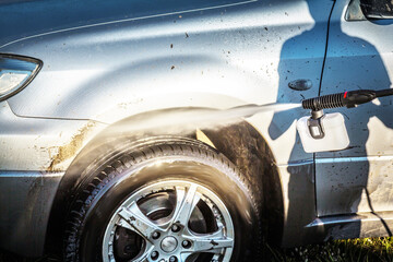 A shadow of a boy with a high pressure washer on a grey dirty car in a sunny summer day.