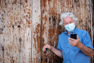 Senior man with white hair using smart phone leaning against a wooden door wearing surgical mask due to coronavirus infection - active elderly people social and technological
