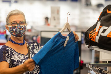 Front view of a senior woman wearing  surgical mask due to coronavirus covid-19 looking a blue tshirt in the store - new normality and consumerism concept