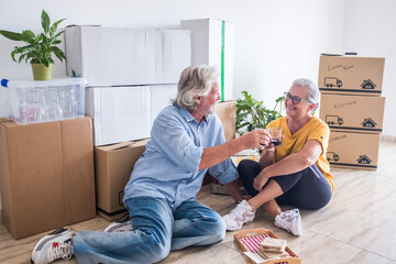 Smiling couple of senior people in the new apartment with moving boxes around them, sitting on the floor toasting with red wine glasses- concept of active elderly people and new beginning like retired