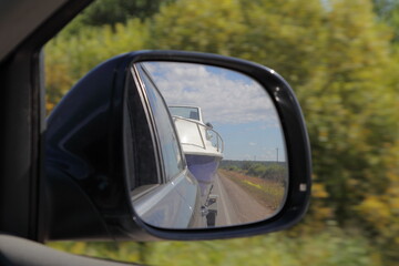 Cabin motor boat towing on car trailer, close up view in reflection in right side car rear mirrow on blue sky and green field background, travel with private boat