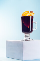 Glass of red glogg or mulled wine with orange slices and cinnamon stick on cube, blue background, vertical