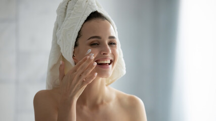 Close up smiling overjoyed young woman with white bath towel on head applying moisturizing face...