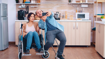 Boyfriend taking selfie with happy and disabled wife in wheelchair. Hopeful husband with...
