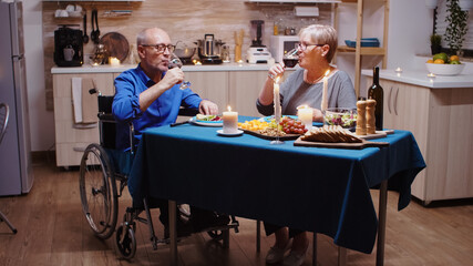 Paralyzed man toasting with wine with his wife. Senior old man in wheelchair dining with aged woman sitting in the kitchen. Imobilized paralyzed handicapped elderly husband having romantic dinner