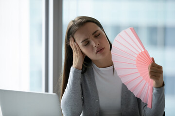 Overheated unhappy young businesswoman waving paper fan in office, sitting at desk with laptop,...