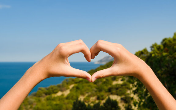 Hands in the shape of a heart against the background of mountains and the sea. Focus on hands, background blurred.