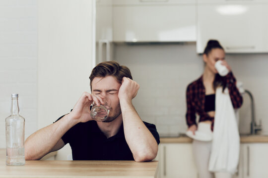 Drinking problem drunk man in a young family. A man drinks while sitting at a table in the kitchen, a woman behind him quarrels and shouts