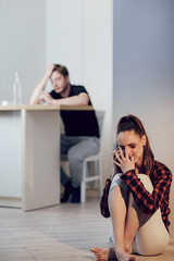 Fototapeta na wymiar Victim of domestic violence. Young scared woman with bruises on face complains to girlfriend on cell phone sitting on the floor in the kitchen. Her husband is sitting behind and drinking alcohol