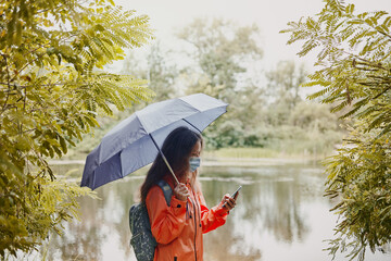Young caucasian woman in a coronavirus mask holding an umbrella using a smartphone in the autumn...