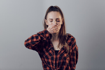 Close up portrait of a frustrated young woman with eyes closed and beaten face holding her hands on the mouth. Woman expressing fear, standing on grey background with copy space