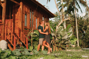 Young lovely couple wearing natural clothes standing near wooden house, hugs and smiles. Honeymoon on tropical island Bali near wooden bungalow