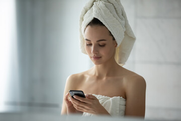Close up head shot young woman wearing white bath towel on head using smartphone, beautiful female...