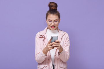 Young pretty European female isolated over lilac background standing in pale pink jacket, texting on cellphone with mate, showing happy friendly smile.