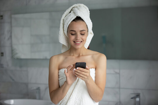 Smiling young woman with white bath towel on head using smartphone after shower, standing in bathroom, happy positive female holding phone, checking social network, reading message, good news - Powered by Adobe