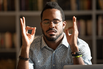 Close up mindful African American man wearing glasses meditating, hands mudra gesture, peaceful calm young male with closed eyes practicing yoga, relieving stress and negative emotions
