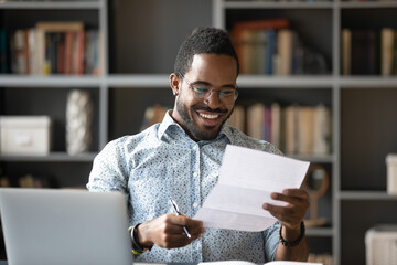 Happy smiling African American man wearing glasses reading letter at workplace, sitting at work...