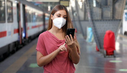 Fototapeta premium Young woman wearing KN95 FFP2 face mask waiting train at the station. Casual woman using smart phone in train station.